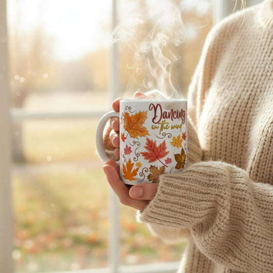 A women wth a cosy knitwear in front of a window holding a white ceramic mug with printed design of warm colored automn leaves with a text saying dancing in the wind.